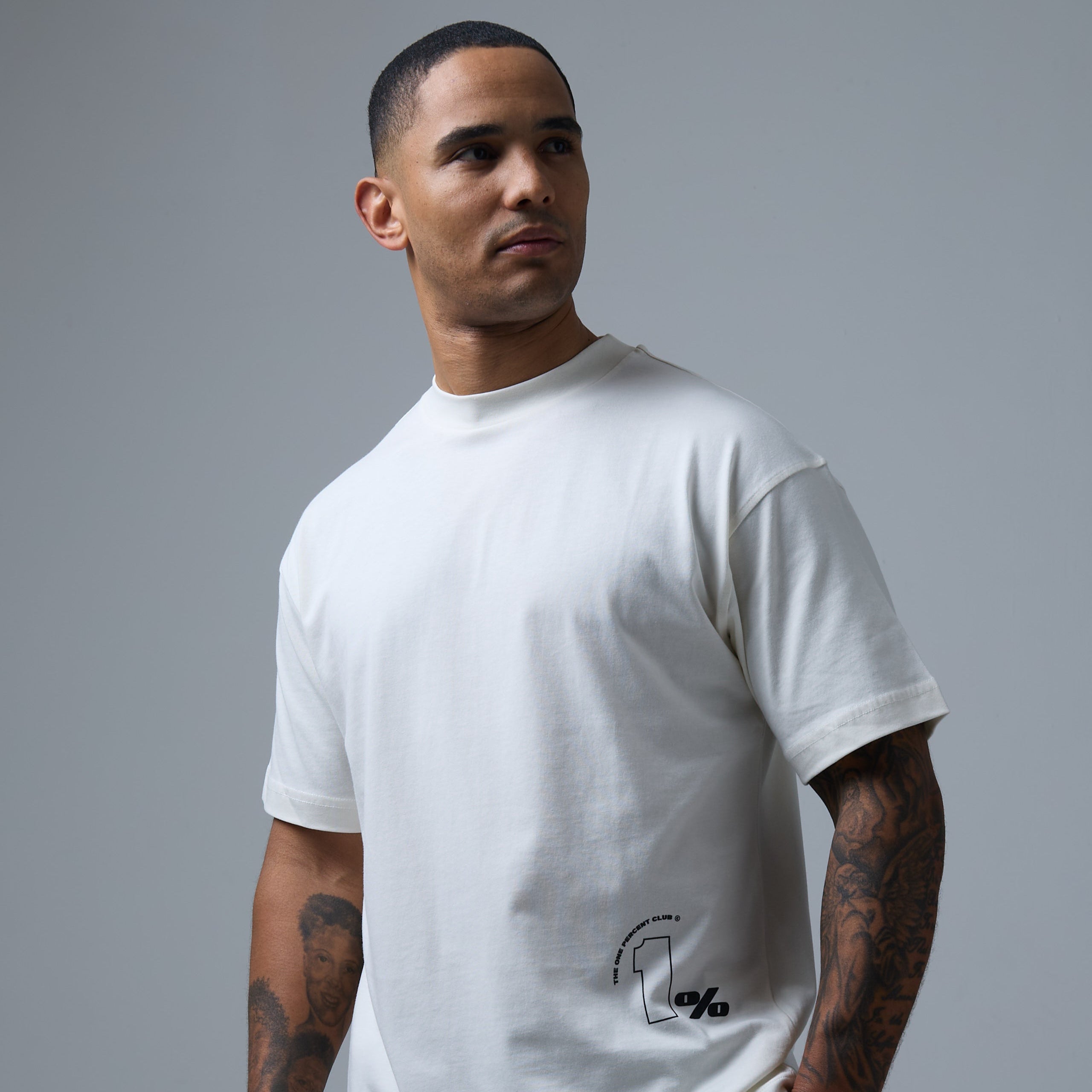 Model wearing a minimalist white t-shirt from The 1% Club clothing brand, featuring the brand's sleek '1%' logo printed near the lower hem. The design emphasises clean lines and exclusivity, aligning with the brand's premium identity.