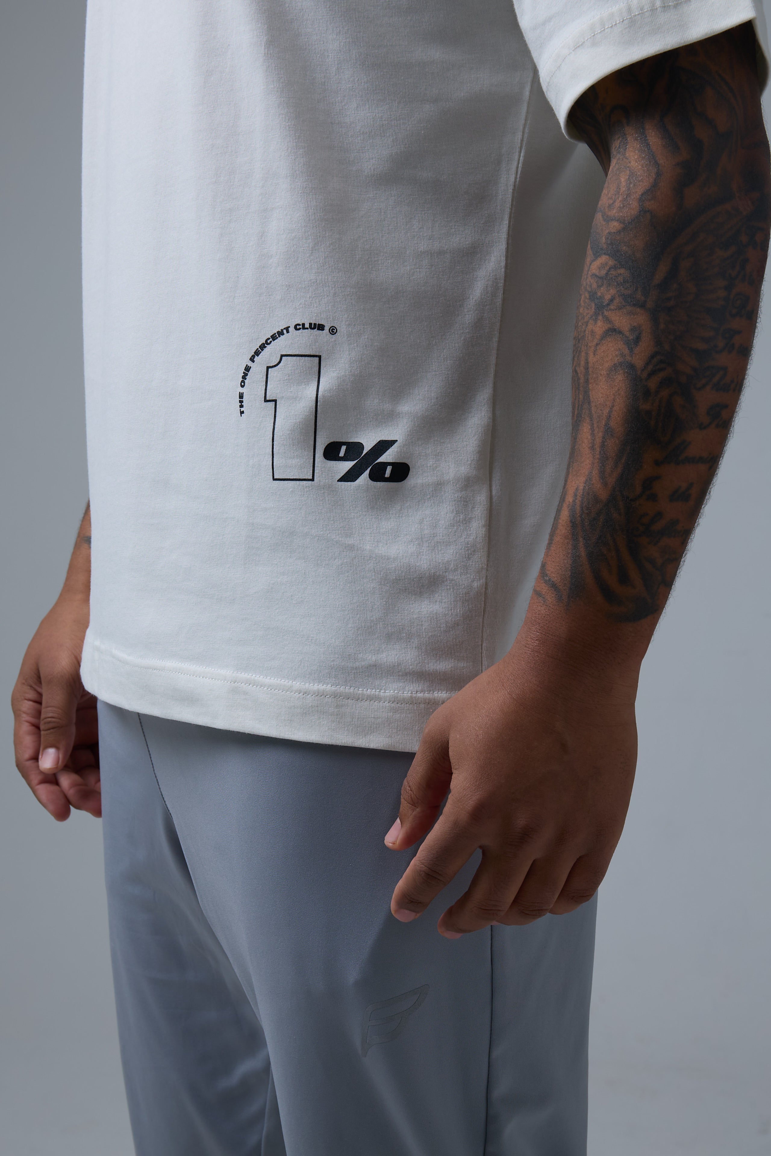 A close-up shot of a man wearing an off-white oversized t-shirt with a minimalist '1% CLUB' logo printed in black on the lower left side. The high-quality fabric drapes smoothly, showcasing a relaxed fit. His tattooed arm, featuring intricate designs, adds an edgy contrast to the clean aesthetic of the shirt. He pairs the t-shirt with light grey athletic joggers, creating a stylish and modern athleisure look. The soft lighting and neutral background highlight the premium details and subtle branding.