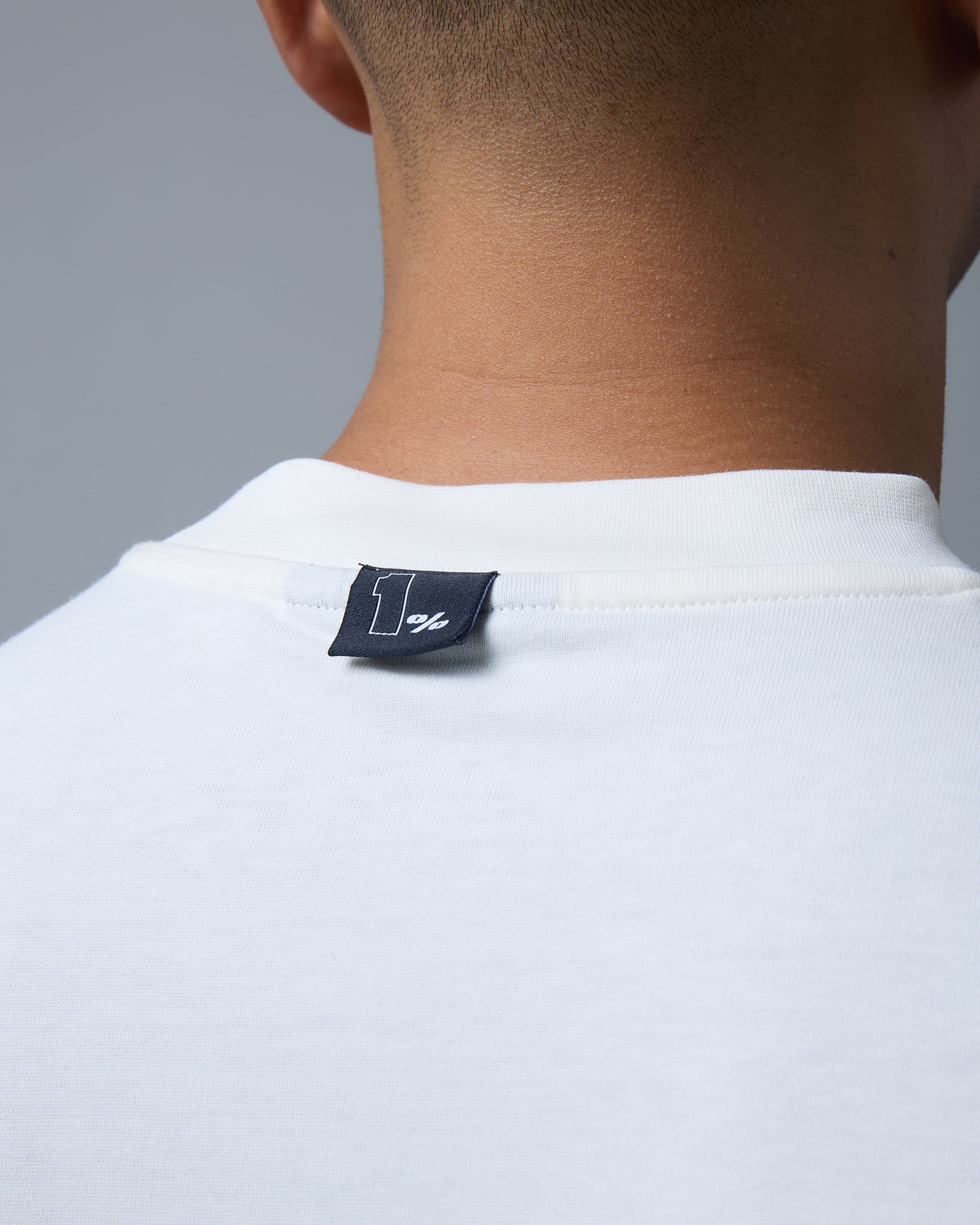 A close-up shot of the back of a man's neck, highlighting the small black woven tag stitched onto the collar of an off-white t-shirt. The tag features a minimalist '1%' logo in white, subtly branding the premium streetwear piece. The finely ribbed neckline and high-quality fabric showcase attention to detail and craftsmanship. The clean, neutral background emphasises the sleek and modern design, making it a stylish and understated addition to contemporary fashion.