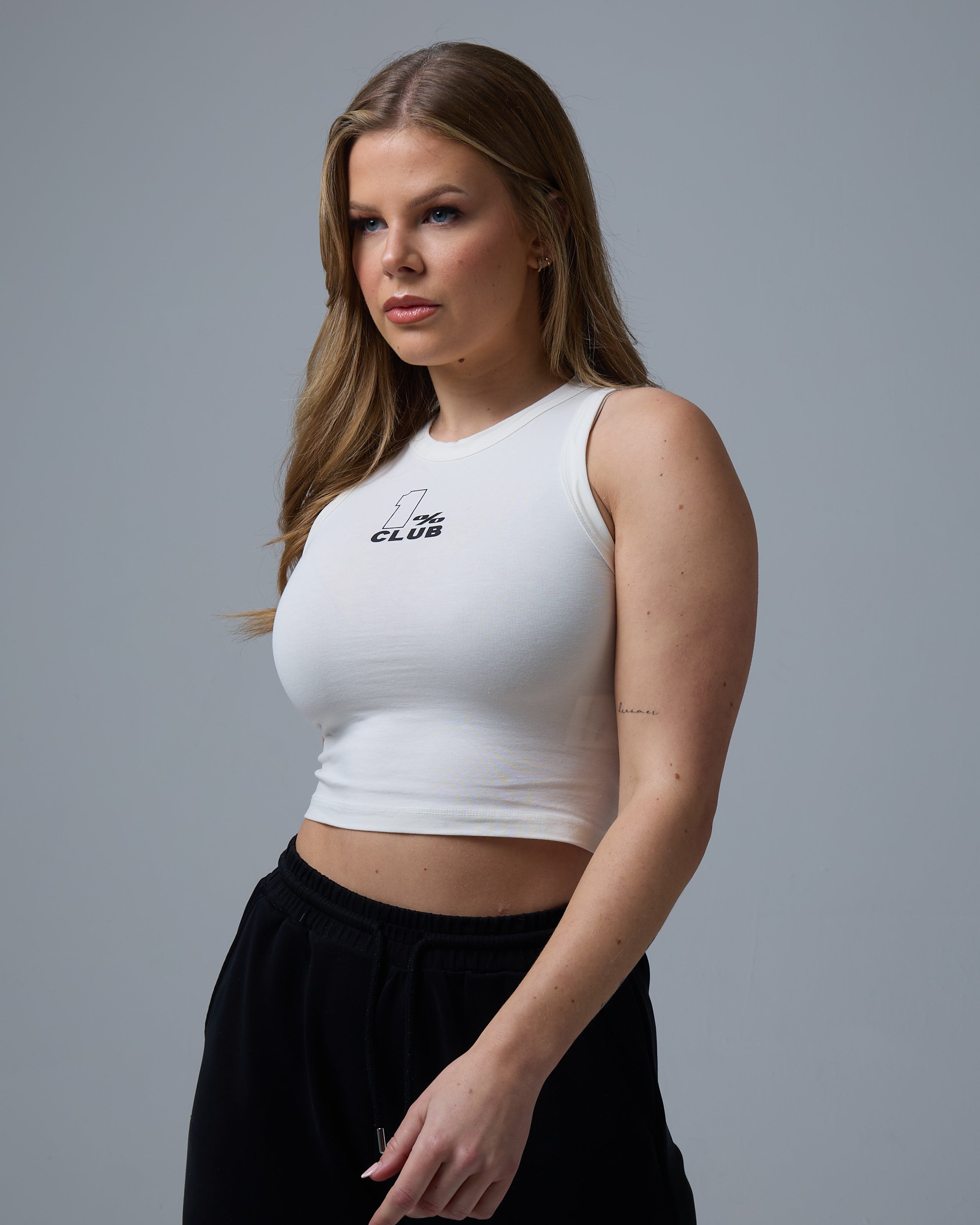 A woman with long, wavy blonde hair poses against a neutral gray background, wearing a off-white cropped racer tee with '1% CLUB' printed in white on the chest. Her arms hang down, showcasing toned arms and a minimalist tattoo on her forearm. She pairs the fitted sleeveless top with black drawstring joggers, creating a sleek and modern athleisure look.