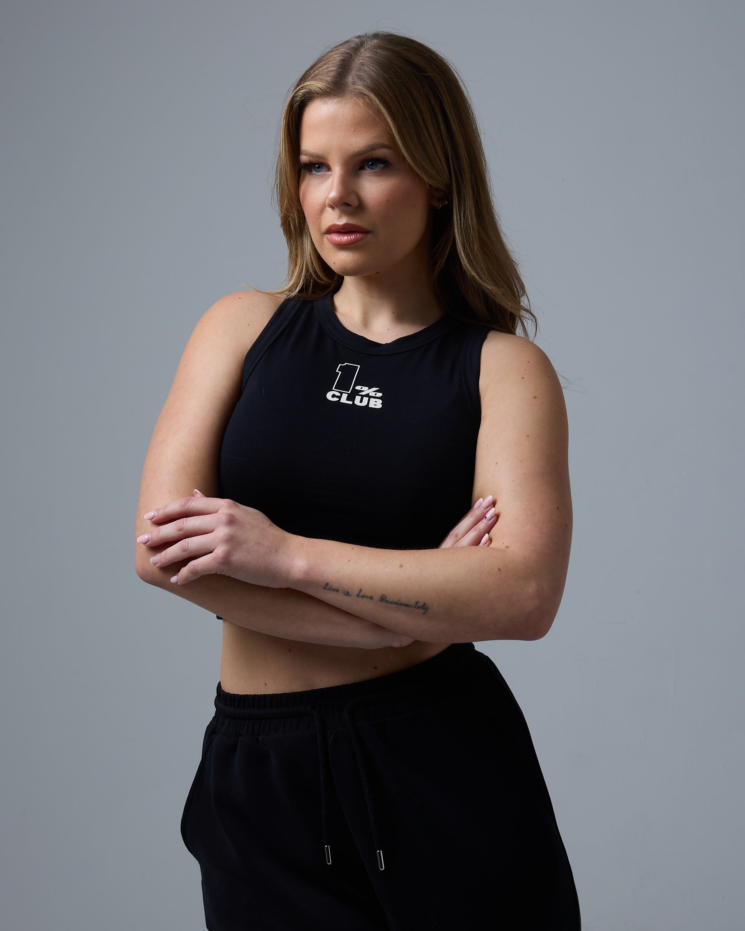 A woman with long, wavy blonde hair poses against a neutral gray background, wearing a black cropped racer tee with '1% CLUB' printed in white on the chest. She crosses her arms, showcasing toned arms and a minimalist tattoo on her forearm. She pairs the fitted sleeveless top with black drawstring joggers, creating a sleek and modern athleisure look.