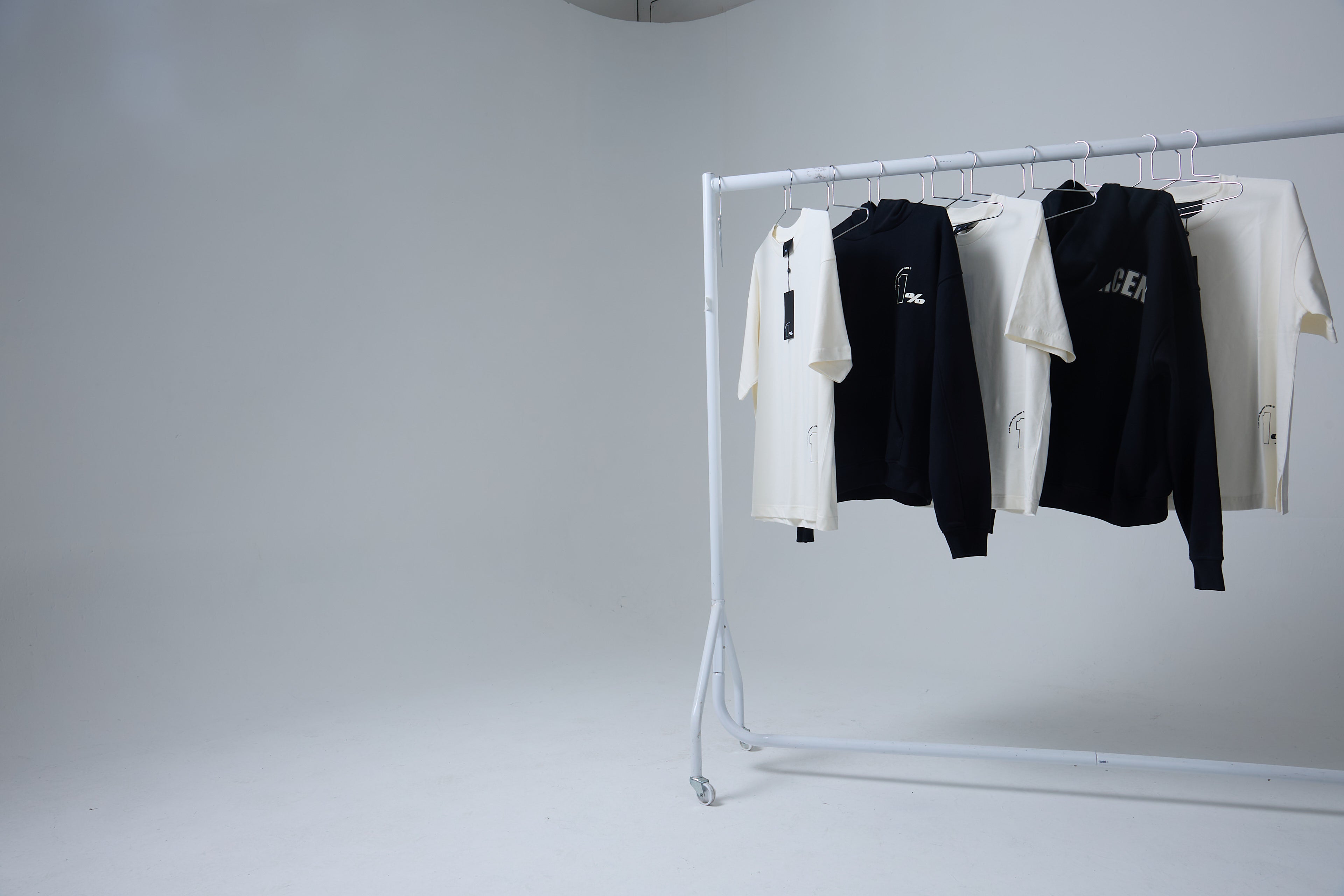 A minimalist clothing rack displaying The 1% Club apparel, featuring a mix of black and white t-shirts and hoodies with subtle branding details. The collection highlights the brand's focus on clean, modern and exclusive designs.