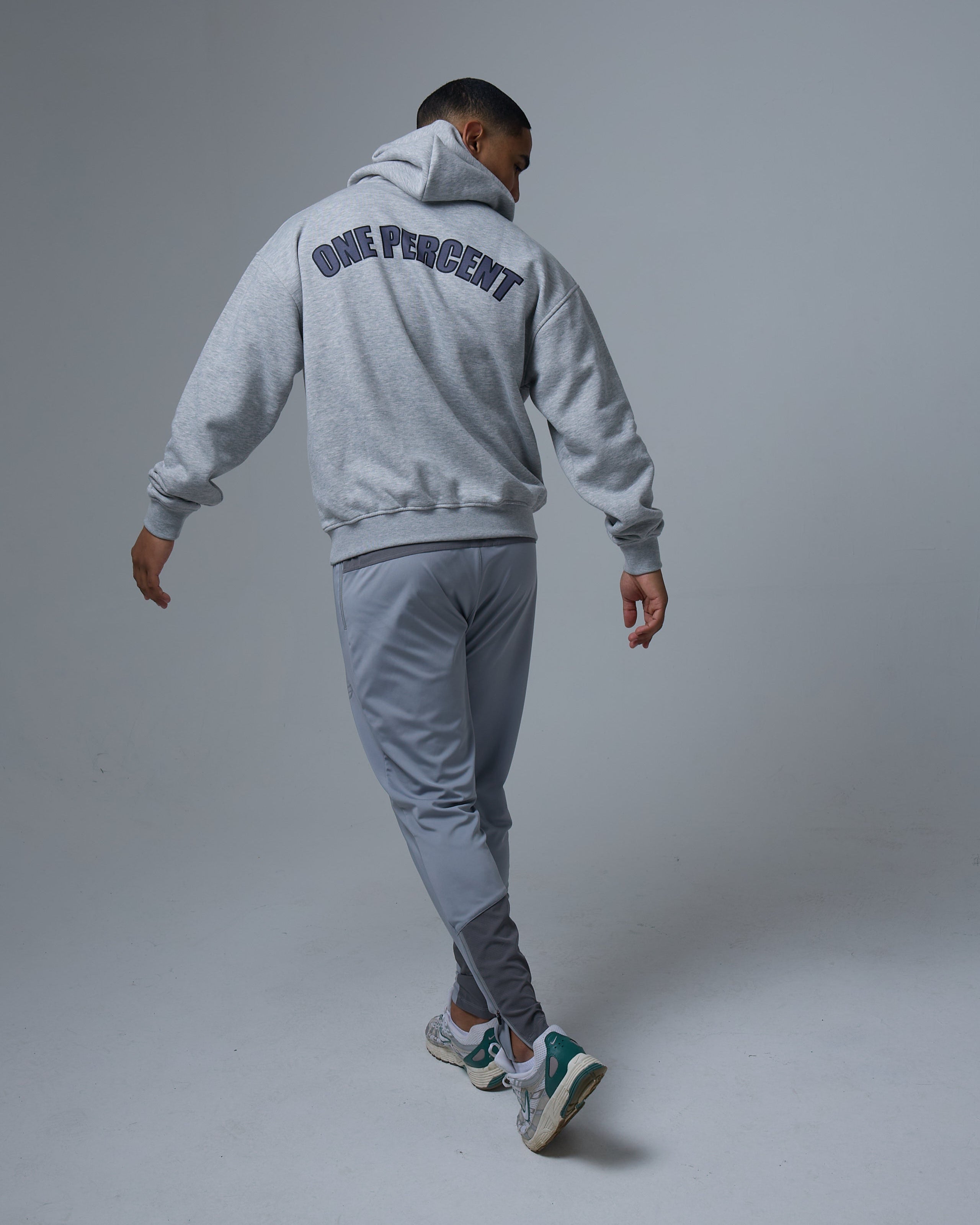 A man walks away from the camera wearing a grey oversized hoodie with 'ONE PERCENT' printed in bold, black capital letters across the back. The hoodie has a relaxed fit, ribbed cuffs, and a spacious hood that is partially up. He pairs it with light grey athletic joggers and stylish sneakers with green and white accents, creating a modern athleisure look.