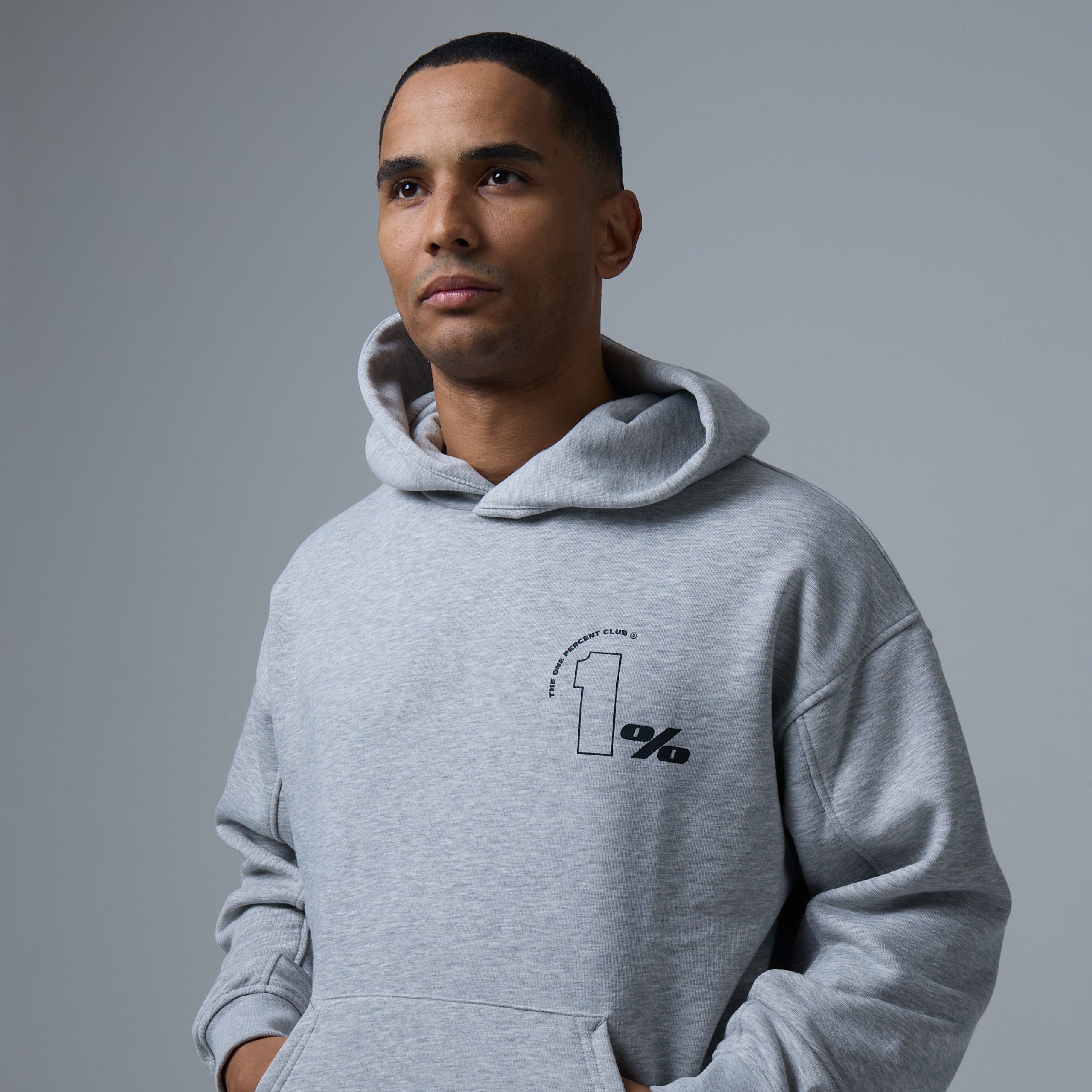 Model wearing a light grey hoodie from The 1% Club clothing brand, featuring the signature '1%' logo on the chest with a circular design element. The hoodie combines casual comfort with a modern, exclusive aesthetic.