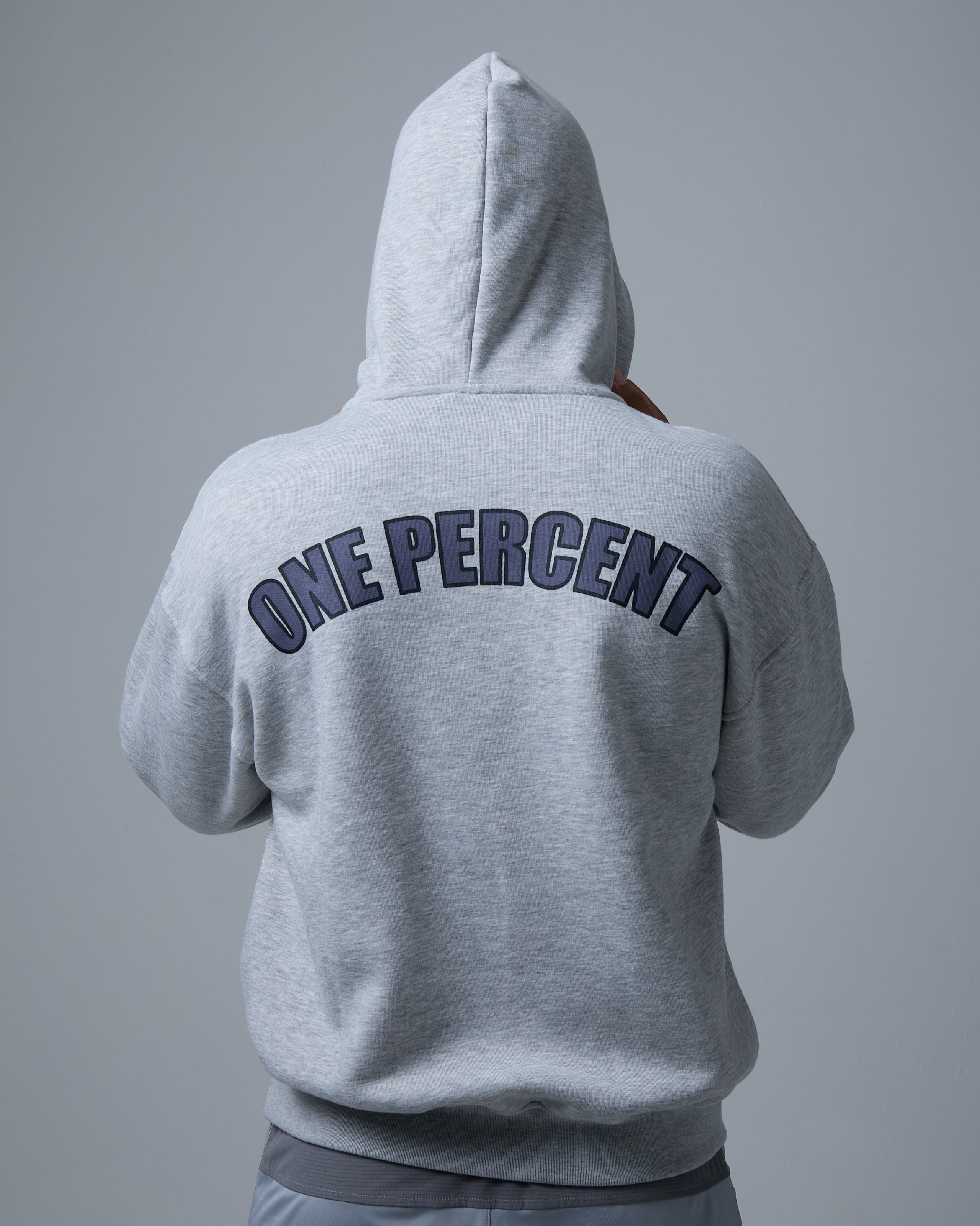 A person stands with their back to the camera, wearing a grey oversized hoodie with 'ONE PERCENT' printed in bold, black capital letters across the back. The hood is up, covering their head, adding a sense of mystery and streetwear appeal. The ribbed cuffs and hem provide a snug fit, while the relaxed silhouette ensures comfort and a modern aesthetic. Paired with light grey athletic joggers, the look embodies a stylish blend of athleisure and casual urban fashion.