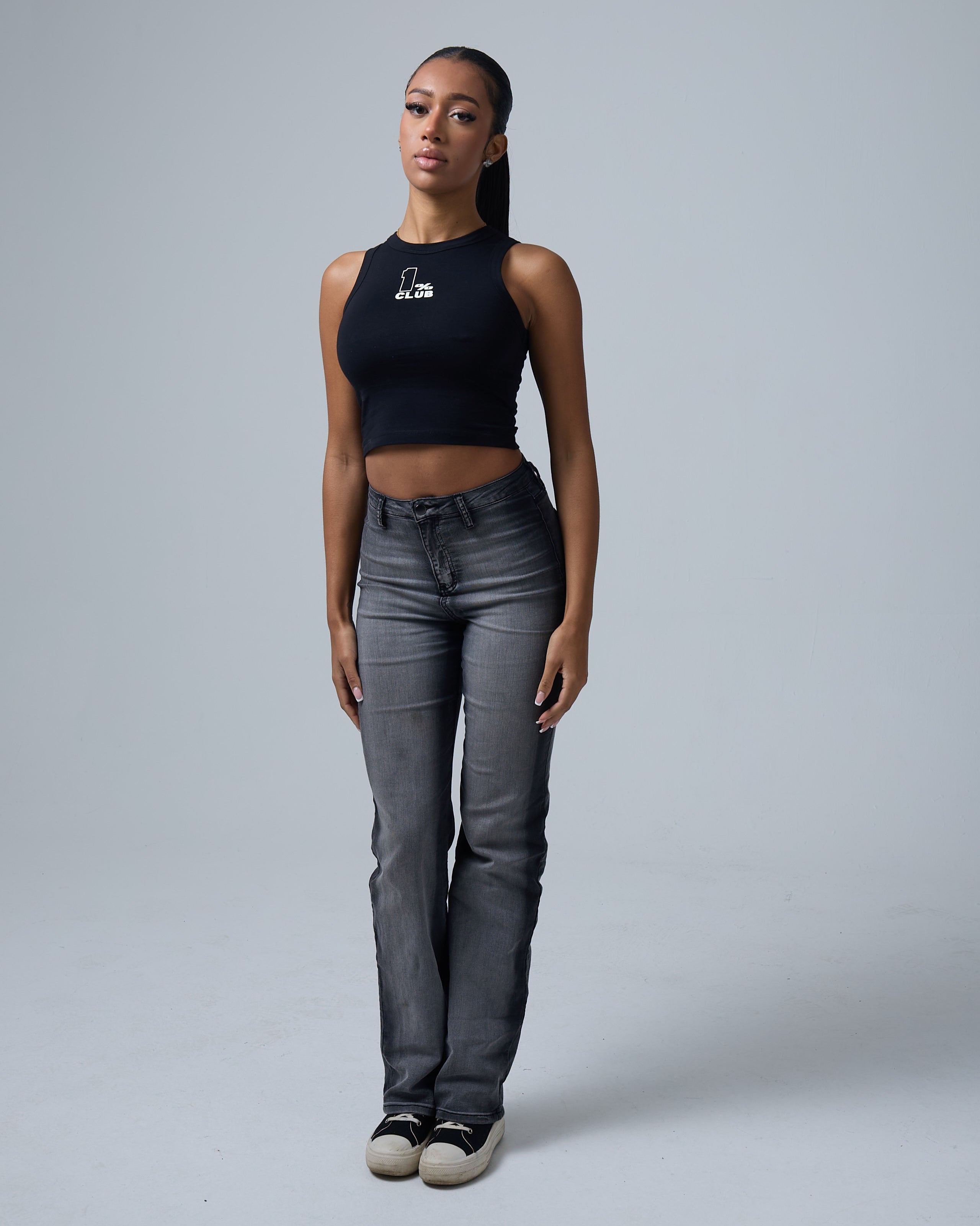 A stylish young woman with a slim, toned physique poses against a neutral gray background, wearing a black cropped tank top with '1% CLUB' printed in white on the chest. She pairs the fitted top with high-waisted, slightly faded gray jeans and black-and-white sneakers, creating a sleek and modern streetwear look. Her hair is pulled back into a sleek ponytail, and she has a confident, relaxed expression. The minimal backdrop and soft lighting highlight the contemporary urban fashion aesthetic.