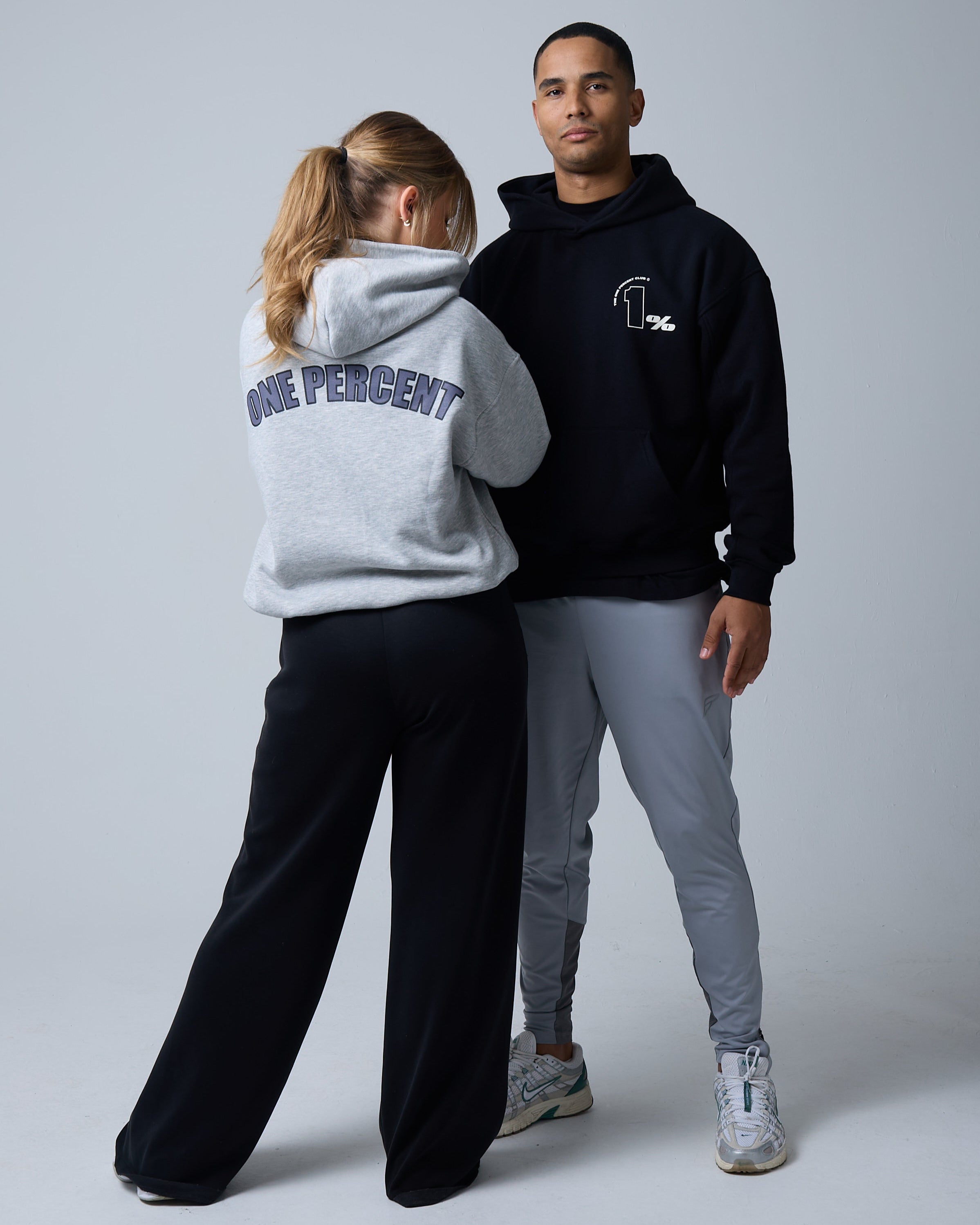 A stylish man and woman pose against a neutral grey background, showcasing modern streetwear hoodies. The woman, facing away from the camera, wears a grey oversized hoodie with 'ONE PERCENT' printed in bold, capitalised letters across the back. She pairs it with loose-fitting black pants and white sneakers. The man, standing confidently beside her, wears a black hoodie with a minimalist '1%' logo on the chest, paired with light grey athletic joggers and trendy sneakers.